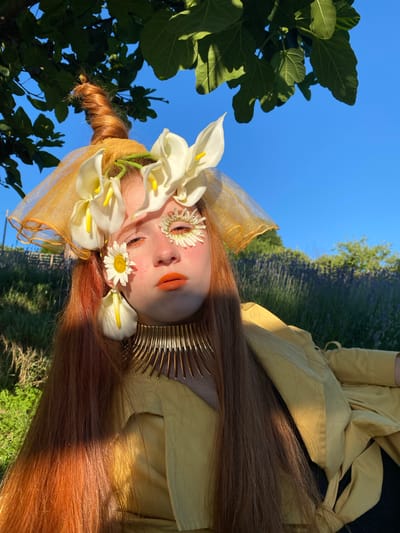 A person with long, reddish-brown hair lies on their back outdoors, their head adorned with white calla lilies and small daisies. Their eyes are closed, and their face is decorated with orange lipstick and flower petals around their eyes. They wear a yellow, draped garment and a metallic gold collar. The background features a bright blue sky, green foliage from a large tree on the left, and a field of purple flowers, possibly lavender, in the distance. Sunlight illuminates the scene, suggesting it is a clear day. The image has been rotated 90 degrees counter-clockwise, so the subjects appear sideways. The overall mood is ethereal and natural.