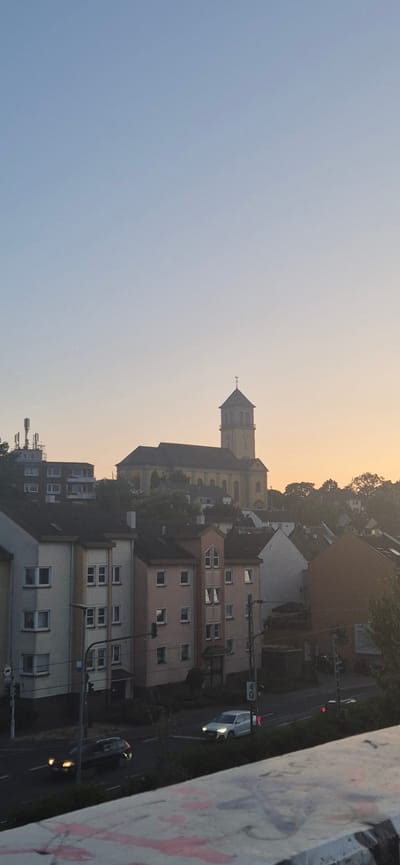 The image captures a view of residential buildings and a church in Mainz, Germany, during what appears to be either sunrise or sunset, given the soft, warm light. In the foreground, a concrete structure with graffiti is visible. Beyond this, a street runs through the scene, with two cars present: a dark sedan on the left and a white SUV on the right. Several multi-story apartment buildings are lined up along the street. In the background, a prominent yellow church with a tall steeple stands on a hill, characteristic of the architecture found in Mainz. The sky above is a pale blue transitioning to a soft orange hue near the horizon, indicating either dawn or dusk. The overall scene depicts a quiet urban environment.