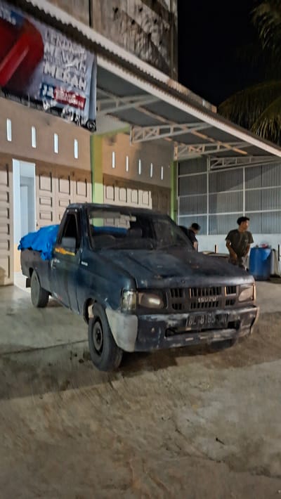 A dark blue Isuzu pickup truck is parked on a concrete surface outside a building at night. The truck bed is covered with a blue tarp. Two men are standing near the back of the truck. The building has a white corrugated metal roof with supporting beams. A banner is hanging on the front of the building. The license plate on the truck reads "BL 8185 JH". The scene appears to be in a public or commercial area in Angkleng Barat, Indonesia.