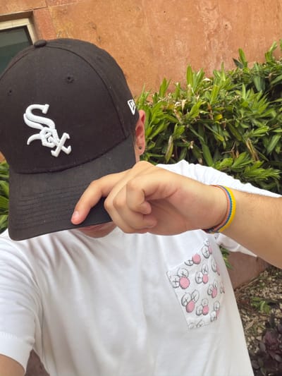 In Mérida, Mexico, a person wearing a white t-shirt with a pocket featuring a pattern of stylized cartoon faces, and a black baseball cap with a white logo, is partially obscuring their face with their hand. On their wrist, a woven bracelet displays bands of yellow, blue, and coral. The background consists of green foliage against a reddish-brown wall.