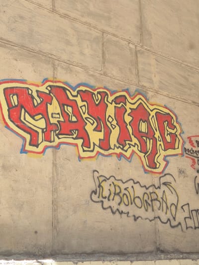 The image depicts graffiti on a concrete wall in Kropyvnytskyi, Ukraine. The primary graffiti is a large, stylized word in red and yellow with a black outline, appearing to be "RANIAS" or a similar variation, rendered in a bubble-letter style. Below this, another line of graffiti in yellow with a dark outline is visible, appearing to read "Kibobrraba" or similar, also in a stylized, wavy font. Further to the right, there is faint dark graffiti and what appears to be a small sticker or piece of paper with writing on it. The concrete wall itself is segmented by vertical lines, suggesting it is made of pre-cast concrete panels. The lower part of the image shows the bottom edge of the wall with some debris or discoloration. The lighting suggests it is daytime.