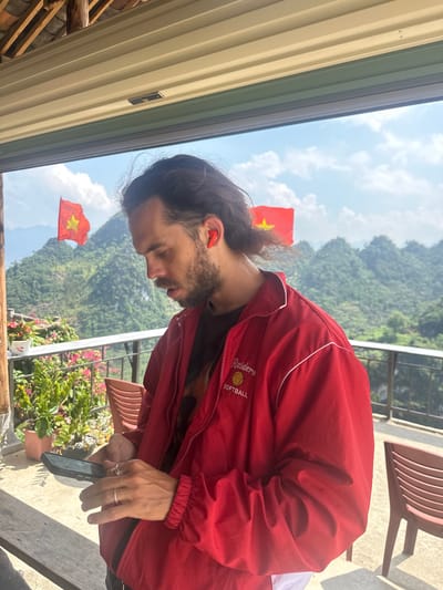 A medium-shot, eye-level view depicts a man outdoors in Lũng Phìn Commune, Vietnam. The man is wearing a red jacket with "Raiders SOFTBALL" embroidered on the chest. He has long, dark hair pulled back and a beard, and he is looking down at a smartphone in his hands. The landscape behind him includes mountains and Vietnamese flags flying in the distance. Floral plants and some seating are visible in the foreground. A portion of a building roof is visible in the upper left corner of the frame.