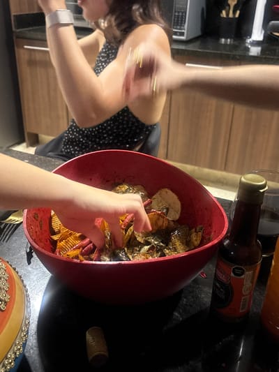 A medium shot captures a red bowl filled with chips and some kind of meat on a black counter. A bottle of salsa inglesa sits beside the bowl. A person's hand reaches into the bowl. Another person wearing a black and white dress is out of focus in the background. A fork is placed near the bottom of the bowl. An item with colors of blue, yellow, and orange is at the lower part of the frame. The location is unknown.