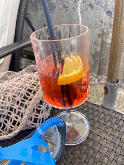 A clear glass filled with an orange-hued beverage sits on a woven outdoor table. The drink is garnished with a slice of orange and appears to contain ice. A dark blue straw is inserted into the glass. To the right of the glass, a light-colored net bag is partially visible, and next to that, a bright blue diving mask with a clear lens. The background shows a blurred outdoor setting with elements that suggest a poolside or beach area, including what looks like gravel and a hint of water. The lighting suggests it is daytime, likely sunny. The overall scene evokes a relaxed, vacation atmosphere. There is no visible text in the image.