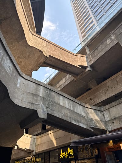 The image shows an upward view of a complex structure, possibly an overpass or elevated roadway, in the Hongkou District of Shanghai, China. The structure is made primarily of concrete, with various supporting beams and columns intersecting at different angles, creating a geometric pattern against the sky.
In the upper left of the frame, a modern building with a glass facade reflects the sky. A sign with Chinese characters is seen to the right side of the frame. The characters are in yellow on a dark background and there is some English underneath the Chinese.