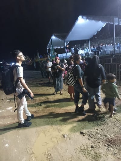 This nocturnal image captures an outdoor gathering or event in Kecamatan Pujut, Indonesia, illuminated by artificial lighting from temporary tents. The dark sky overhead confirms it is nighttime.
In the foreground, several individuals are standing on uneven, muddy, and dusty ground. To the left, a young man with short dark hair and glasses wears a striped t-shirt, light trousers, and dark shoes, with a backpack on his shoulders and a camera in his right hand. He looks upwards and to the left, as if observing something outside the frame.
Towards the center and right, other people are visible. A man with a red head covering and a dark t-shirt appears to be holding a camera, standing near another woman wearing a black and white striped backpack and dark clothing. Further right, a woman in a dark hoodie and light pants holds the hand of a small child. The ground beneath them shows patches of muddy water and rough terrain.
In the background, under brightly lit white canopies, an audience is seated in rows of white chairs, suggesting a formal or semi-formal event is taking place. A green flag or banner is partially visible near one of the tents. Metal fencing or barriers separate the foreground area from the seated attendees. The overall atmosphere suggests a community gathering or performance illuminated for the evening. No discernible text is visible on any banners or signs.