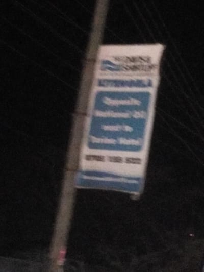 A white and blue sign is affixed to a pole in the dark. The sign reads "Wine-Sense Sommelier," followed by "Experience the difference, certified & Insured by Torbaine-Walter." The bottom of the sign displays the phone number "8782-1283-6322." The sign appears faded or blurry. Wires are visible in the background.