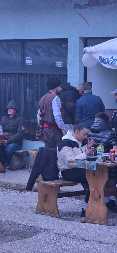 The image shows a group of people gathered outside a building, possibly in Breze, Bulgaria. The group is sitting at a wooden table. There is a man in traditional Bulgarian clothing with a brown vest and embroidered sleeves, seated amongst the group. A woman sits next to him using a mobile phone. There are several cans of drinks on the table. Another man in a hooded jacket is visible seated on the ground in front of the building, resting his head. The building's facade is painted in a light blue tone and has a poster on the wall. A white banner hangs in the top left of the shot. The ground is a grey-colored, grainy material.