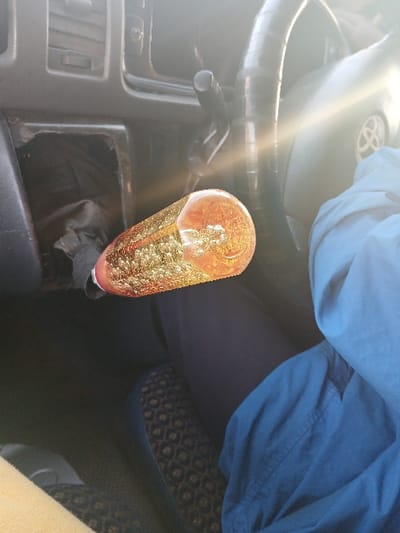 The image depicts the interior of a vehicle, likely a car or van, under strong sunlight. The view is from a low angle, encompassing portions of the dashboard, steering wheel, and the lower bodies of at least two occupants.
**People:** Two individuals are partially visible. On the right, a person's legs are present, clad in dark blue trousers, with a light blue shirt or jacket draped over their lap. The seat beneath them features a dark, patterned fabric. On the far left, a portion of another person's leg or lower body, covered in light-colored fabric, is visible. No faces or upper torsos are discernible.
**Subjects/Objects:**
* **Gear Shift Knob:** Centrally prominent, an elongated, cylindrical gear shift knob made of clear or amber-colored material is visible. It contains a shimmering, golden-orange substance, possibly glitter or liquid.
* **Dashboard:** On the left, a dark gray dashboard features air vents. Below these, an empty, rectangular cavity is present, revealing dark fabric or material within, consistent with a missing radio unit or storage compartment.
* **Steering Wheel:** Partially visible on the right, a dark steering wheel features a central emblem resembling a Toyota logo.
* **Sunlight:** A strong lens flare originates from the upper right, indicating direct sunlight entering the vehicle.
**Actions/Interactions:** No discernible actions or interactions are depicted.
**Environment:** The scene is confined to the interior of a vehicle, with evidence of bright external lighting. The specific location within Thika Township ward, Kenya, cannot be determined from the visual information provided in the image.