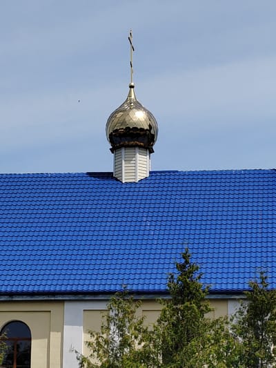 The image shows the golden dome of a church in Воўчынскі сельскі Савет, Belarus. The dome is topped with a cross and sits atop a white structure that protrudes from a vibrant blue tiled roof. The roof covers a building with a light yellow facade, partially obscured by the foliage of green trees in the foreground. The sky is a clear blue with some wispy clouds.