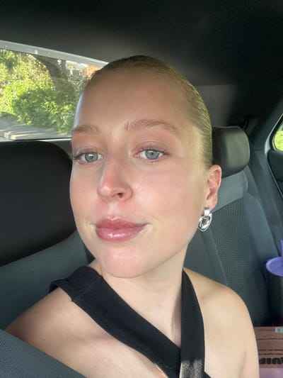 A young person with short blonde hair and light skin is in the driver's seat of a car. The person is wearing a black tank top and a silver earring in their left ear. The car seat is dark gray. Outside the window, green trees are visible. The location cannot be determined from the image.