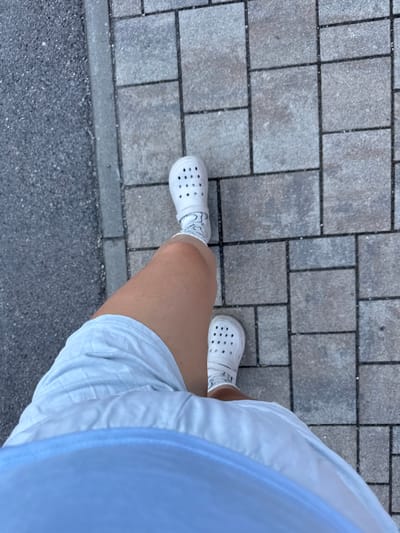 The image shows a person walking on a paved sidewalk. The sidewalk is made of gray rectangular and square pavers arranged in a pattern. To the left of the sidewalk is a dark gray asphalt surface, possibly a road or path. The person is wearing white Crocs and white socks with a subtle pattern. They are also wearing light blue shorts and a light blue top, only the lower portion of which is visible. The shot is taken from a high angle, looking down at the person's legs and feet. The scene appears to be outdoors, likely in an urban or suburban environment. The location is specified as Höhenkirchen-Siegertsbrunn, Germany.