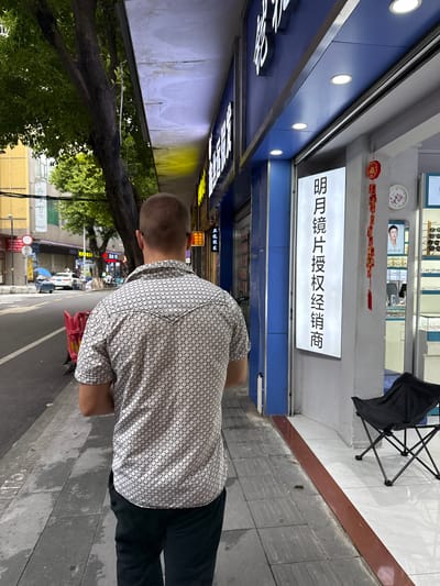 A man walks down a sidewalk in front of a storefront. He is wearing a short-sleeved patterned shirt and dark pants. The storefront has a blue facade and a large white sign with Chinese characters. To the right of the sign is a display of eyeglasses. A folding chair is placed outside the store. The sidewalk is made of concrete tiles, and there is a street to the left of the sidewalk with cars and trees. The location is in Liwan District, China.