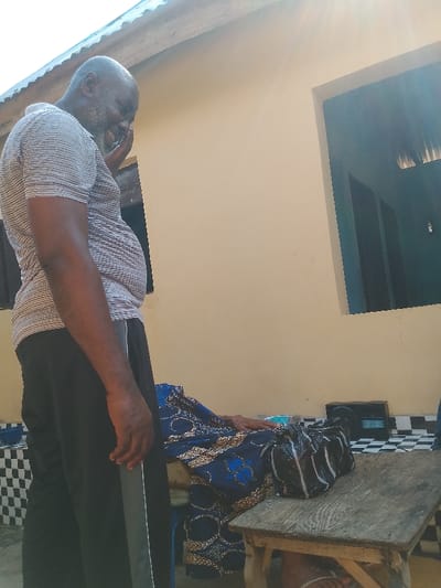 The scene depicts an outdoor setting in Ajalli, Nigeria, characterized by a single-story building with a light beige wall and a corrugated metal roof. The ground is covered with black and white checkerboard tiles. A man with dark skin, a beard, and a receding hairline stands on the left, wearing a striped polo shirt and dark pants with a grey stripe. He is angled slightly to the right, looking downwards, and appears to be smiling or laughing, with his right hand near his face.
To the right and below the man, a second individual is partially visible, seated or reclined on a low surface. This person wears a blue patterned fabric, and one hand rests on their lap. A small, weathered wooden table is positioned in the foreground, holding a black plastic bag with white markings. Behind the table, a black electronic device, possibly a radio, sits on the tiled floor. A foot wearing a white sandal is visible beneath the table. Strong sunlight illuminates the scene from the upper right.
