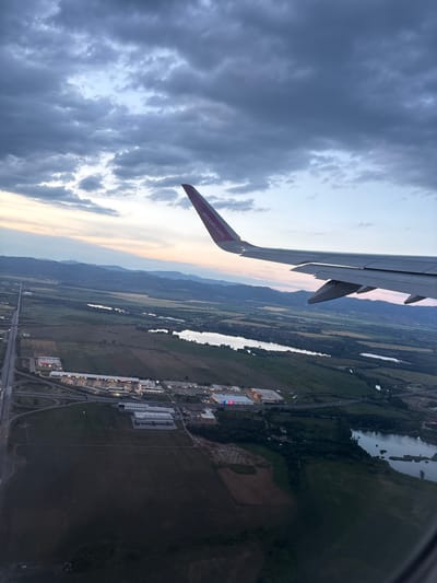 The image, taken from an airplane window, captures an aerial view of the outskirts of Krivina, Bulgaria, during a twilight hour. The scene is dominated by a vast expanse of sky filled with dark, textured clouds, with hints of pale pink and orange light peeking through near the horizon, suggesting either sunrise or sunset.

In the foreground, the wing of the Wizz Air airplane is prominently visible, extending from the right side of the frame. The wing is gray and metallic, with flaps and ailerons clearly discernible. The Wizz Air logo and website address are visible in purple lettering on the tip of the wing.

Below the wing, a sprawling landscape unfolds. A highway or major road runs vertically from the top left of the frame, curving into an interchange with multiple on- and off-ramps. Adjacent to the road, a commercial area is identifiable, featuring several large buildings with illuminated windows, possibly shopping centers or industrial facilities. Some of these buildings have colorful lighting, suggesting active businesses.

Further into the landscape, various fields and agricultural areas are visible, delineated by changes in color and texture. Several bodies of water, likely lakes or ponds, are scattered throughout the scene, reflecting the dim light of the sky. In the distance, a range of mountains forms a hazy backdrop, their silhouettes softened by the atmospheric conditions. The overall impression is one of a developing urban edge meeting rural land, viewed from a considerable altitude.