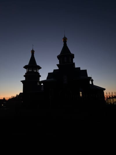 The image displays the Church of the Nativity of the Blessed Virgin Mary (Церковь Рождества Пресвятой Богородицы) in Новинки, Nizhny Novgorod Oblast, Russia, presented as a dark silhouette against a twilight sky. The church features two primary structures: a main building with a prominent golden onion dome topped by an Orthodox cross, and an adjoining bell tower to its left, capped with a smaller golden dome and a spire. The architectural details, including multiple tiers and window outlines, are visible despite the deep shadow. A single interior light source illuminates a window on the right side of the main church building. The sky exhibits a gradient, transitioning from a deep indigo at the top to a faint orange and pink hue along the horizon line on the left, indicative of either late dusk or early dawn. The foreground is completely dark, obscuring any ground-level details. Blurred outlines of distant trees or structures are faintly visible on the horizon to both the left and right sides of the church. No people or specific actions are discernible.