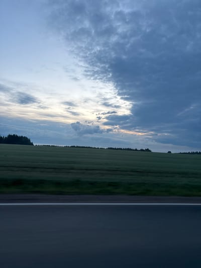 The image shows a rural scene taken from a road. The sky is filled with grey and white clouds. A field stretches towards the horizon. A line of trees separates the field from the sky. To the right, a dark paved road with a white dividing line is visible. The location is near Perevoznoye, Russia. No people or objects are clearly identifiable in this image. The time of day appears to be dusk or dawn.