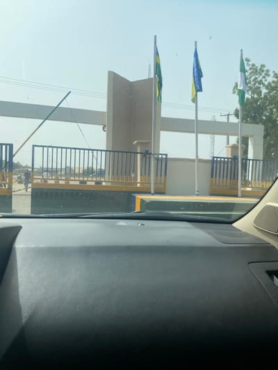 The image depicts the entrance gate of the Nigeria Police Academy in Yan Dala, Nigeria, viewed from the interior of a vehicle through its windshield. The lower portion of the frame is occupied by the dark grey dashboard.

The entrance consists of a substantial light-colored concrete architectural structure forming an archway or gate. In front of this structure are three flagpoles. From left to right, the flags displayed are: a green and yellow flag, a blue flag with "NIGERIA POLICE ACADEMY" text in yellow, and the national flag of Nigeria (green, white, green).

A black metal barrier gate with vertical bars, featuring yellow and blue details on its lower sections, is visible across the entrance. A yellow and black striped boom barrier arm is positioned on the left, angled upwards, indicating an open state. The ground features concrete curbs painted in yellow, blue, and green.

Beyond the open gate on the left, two indistinct male figures are visible; one appears to be walking away from the gate, while the other stands near the roadside. The background reveals a clear, bright sky, with utility lines crossing horizontally. Sparse trees are visible on the far right, and distant structures are faintly discernible.