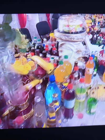 The image depicts a close-up, slightly blurry view of a table laden with various food items and beverages, suggesting a celebratory or communal gathering. The items include a tiered cake under a clear plastic dome, several bottles of colorful drinks (some appearing to be soda or juice), bananas, grapes, and other fruits. The arrangement suggests a buffet or a spread of offerings. The background shows hints of red and white fabric, possibly decorations, and dark chairs, implying an indoor setting. The overall impression is one of abundance and festivity. The image's context in Pavlohrad, Ukraine, adds a layer of specific geographical and cultural information, though this is not visually evident in the provided frame.
