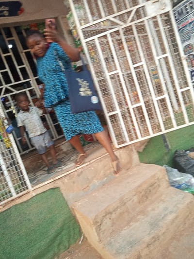 The image shows a woman in a blue and black patterned dress and sandals, carrying a dark blue bag, exiting a storefront. She is walking down a set of concrete steps. A young boy in a plaid shirt and grey shorts is standing on the top step to the right. The storefront is secured with a white metal grate, with products visible inside. The storefront has a concrete exterior. Green artificial grass covers the ground in front of the store. The location of the scene is Ota, Nigeria, based on the provided coordinates. The time of day is indeterminate.