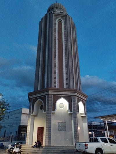 A tall, octagonal minaret with a dome is the central focus of the image, standing against a twilight sky with scattered clouds. The minaret features vertical bands of alternating brick and gray material, with arched decorative elements near its base. A white pickup truck is parked to the right of the minaret, and a motorcycle is visible to the left. Two individuals are seated on the steps leading up to the minaret's entrance, which has a red door. In the background, a building with a flat roof and multiple windows is visible. The scene appears to be in an urban or semi-urban environment, with power lines strung across the upper portion of the image. The overall lighting suggests it is either dusk or dawn.