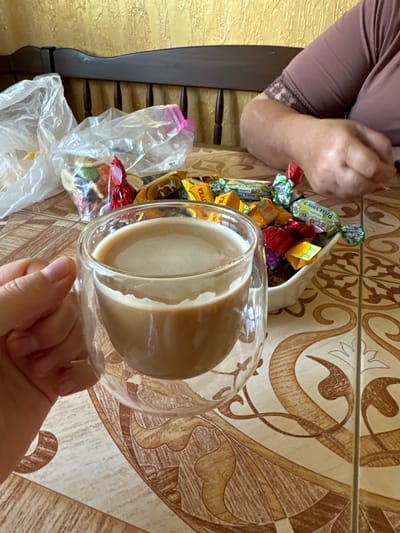 A close-up shot shows a person's hand holding a transparent mug filled with a light brown beverage, likely coffee with milk. The mug has a double-walled design. In the background, a bowl of assorted wrapped candies is visible, with some candies spilling out onto a patterned tablecloth. To the left of the candies, plastic bags are present, one of which appears to be a ziplock bag. A person's arm and shoulder, clad in a dark purple garment with lace trim, are partially visible in the upper right quadrant of the image. The environment suggests an indoor setting, potentially a dining area, given the presence of the tablecloth and the furniture in the background. The overall scene depicts a moment of relaxation, possibly a coffee break with sweets. The image is dated to July 2023, and the metadata suggests it was taken in Chemerne, Ukraine.