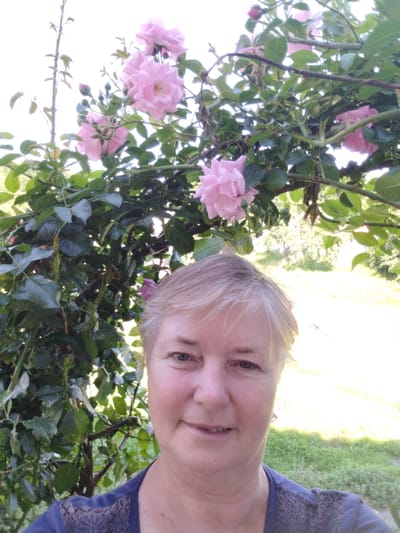 A medium shot captures a woman with short, blonde hair and fair skin smiling directly at the camera. She is positioned in front of a rose archway adorned with clusters of light pink roses and lush green leaves. The background reveals a sunlit garden with hints of trees and grass, suggesting an outdoor setting in Novoselivka, Ukraine. She is wearing a dark blue t-shirt with lace detailing on the shoulders and neckline. The overall lighting is bright and natural, indicating it was taken during the daytime.