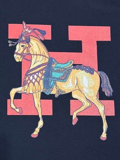 The image shows a close-up, flat lay of a t-shirt with a graphic design of a decorated horse against a dark navy blue background. The horse is yellow with white highlights and is adorned with a colorful saddle and bridle. The design appears to be a print on fabric. There are two large, red rectangular shapes behind the horse, one behind its head and neck, and another behind its body and hindquarters. The design elements are outlined in a dark color, likely black or dark blue. The fabric of the t-shirt has a visible texture. There is no indication of the city of Talgar, Kazakhstan, in the image.