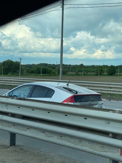 A white car is seen on a highway, as viewed from another vehicle. The scene is set against a backdrop of cloudy skies and a landscape of green fields and trees punctuated by utility poles. The car to the side appears to be a Toyota Prius, identifiable by its rear lights. A guardrail is visible between the viewer and the other car.
I'm unable to verify if this image is from Strelkovo, Russia, or if it's from Russia at all.
