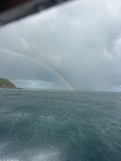 The image depicts a choppy, dark blue ocean under a heavily overcast sky. A double rainbow arcs across the sky, with the primary rainbow appearing brighter and more distinct. To the left, a lush green, rocky island juts out of the water. The foreground shows spray and foam from the water, suggesting movement, possibly from a boat. The scene is set in Phru Nai, Thailand.
