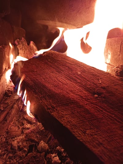 This media file depicts a close-up view of a fire burning in a fireplace. The primary subjects are logs of wood, embers, and flames. The scene is set within the confines of a fireplace, suggested by the brickwork visible on the right side of the image and the ash and glowing coals on the bottom.

The activity taking place is the burning of wood, indicated by the active flames licking around a large log in the foreground and smaller pieces of wood in the background. The flames are a mix of bright orange, yellow, and hints of blue near the base of the fire, suggesting intense heat. The log in the foreground is reddish-brown with a textured, grainy surface and appears to be partially consumed by the fire. Ash and glowing embers are scattered around the base of the burning wood, creating a warm and textured landscape.

Notable details include the intimate and warm glow cast by the fire, which illuminates the scene. The time of day is not explicitly indicated, but the warmth and glow suggest an indoor setting, possibly during cooler hours. There are no visible signs of weather or specific location cues beyond it being a fireplace. The overall mood conveyed is one of warmth and coziness. No text is visible in the image. The provided location context of Poltavka, Kyrgyzstan, suggests the setting is likely a home or dwelling in that region.