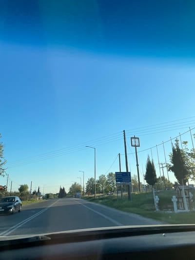 The image captures a bright, sunny day along a road in the Xanthi Municipal Unit, Greece, viewed from inside a moving or stationary vehicle. The clear blue sky dominates the upper portion of the frame, crisscrossed by numerous overhead power lines connected to wooden utility poles and metal streetlamp posts.

On the right side of the road, a stretch of green grass runs parallel to the asphalt, backed by a line of trees. Partially visible beyond the trees is a white stone structure or fence, suggesting a cemetery or a significant roadside feature. Further down the road, more trees and hints of buildings can be seen under the bright sun.

A prominent blue directional sign is visible on the right, providing navigational information. The text on the sign, in Greek, reads: "ΚΑΒΑΛΑ 51" (Kavala 51 km), accompanied by a left-pointing arrow, and "ΚΟΜΟΤΗΝΗ 150" (Komotini 150 km), with a straight-ahead arrow. This indicates distances to these cities, confirming the regional location in Northern Greece.

In the foreground, a black car is visible on the two-lane asphalt road, which features clear white lane markings. The time of day is clearly mid-day, with strong sunlight and no clouds in the sky, contributing to a sense of a pleasant driving day.