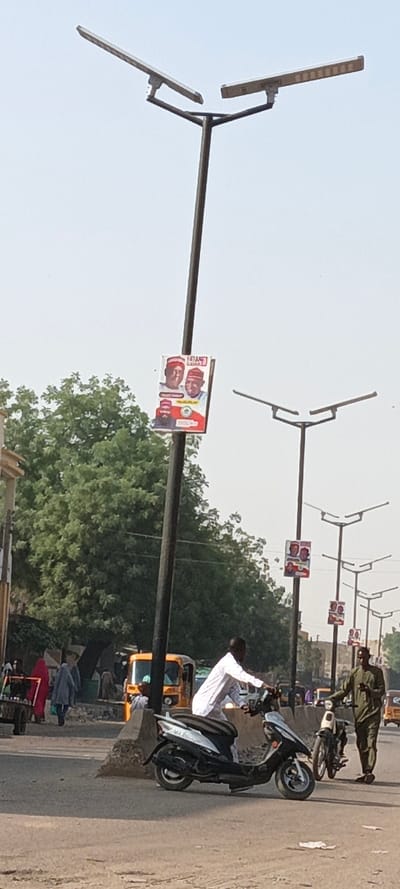 This image captures a daytime street scene in Kaura Goje, Nigeria. A dark-colored streetlight pole with a bifurcated top supporting two rectangular solar panel/LED light fixtures dominates the left-center foreground. A rectangular political campaign poster, featuring three male individuals in traditional attire and red caps, is affixed to this pole. To the right, a series of identical solar-powered streetlights extends into the distance, each displaying similar campaign posters.
On the paved road surface in the mid-ground, a man dressed in a white tunic is situated on a white and black scooter, facing right. To his immediate right, another man in an olive-green traditional outfit stands next to a lighter-colored motorcycle. In the background on the left, a large, dense green tree is visible. Several pedestrians, including individuals in red and light blue garments, are walking along the side of the road. Multiple yellow auto-rickshaws (Keke Napep) are also present further down the street. The road surface exhibits signs of use, and a concrete barrier is visible at the base of the main streetlight pole. The sky is clear and bright.