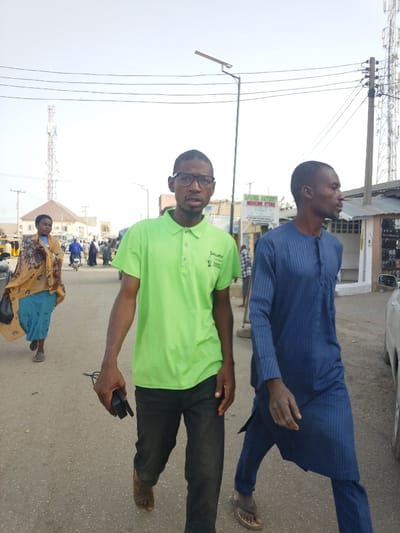 The scene is an outdoor street in Garun Gawa, Nigeria, under bright daylight. In the foreground, two adult males are walking. The man on the left wears a lime green polo shirt, dark trousers, and black-rimmed glasses; he holds a black electronic device in his right hand and looks directly at the camera. The man on the right wears a dark blue striped long-sleeved tunic and matching trousers, walking to the right with his gaze directed forward. In the midground on the left, an adult female in a light blue dress and a patterned yellow and brown head covering walks away from the camera. The background depicts an urban environment with an asphalt road, several pedestrians, and motorbikes. Structures visible include a one-story building with a red-brown roof on the left, and a white building with a corrugated metal roof featuring a sign that reads "APUNA PATIENT MEDICINE STORE" on the right. Overhead, utility lines traverse the frame, and a solar-powered street light is visible. Two lattice telecommunication towers are present in the distant background, one on the left and another on the far right.