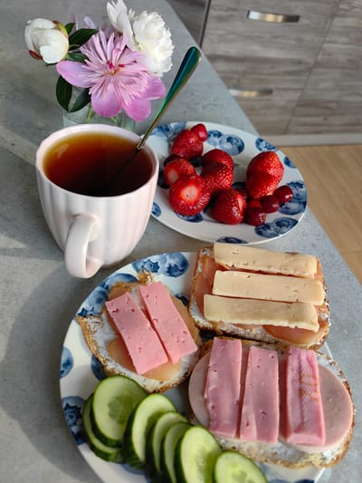 The image shows a breakfast scene on a countertop, likely in a kitchen, with a hint of wooden cabinets in the background. In the foreground, on a grey speckled countertop, there are two plates of food. The plate on the left contains sliced cucumber and two open-faced sandwiches. One sandwich has a pink, processed meat slice topped with two rectangular pieces of pink cheese. The other sandwich has a round slice of processed meat topped with three rectangular pieces of pink cheese. The plate on the right has a cluster of strawberries and cherries, and another open-faced sandwich topped with three rectangular slices of pale yellow cheese, possibly aged Gouda, and thin slices of ham.
To the left of the plates, a light pink mug filled with dark tea is present, with a spoon inside. Beside the mug, a small glass vase holds a bouquet of peonies and other flowers, with some in full bloom and others as buds. The lighting suggests it is daytime, with shadows cast across the countertop.
While the image depicts a breakfast setting with food and flowers, there is no indication or visual information within the image that specifically places this scene in the city of Fanipal, Belarus. The objects and environment are common and do not possess any unique identifiers related to that location.