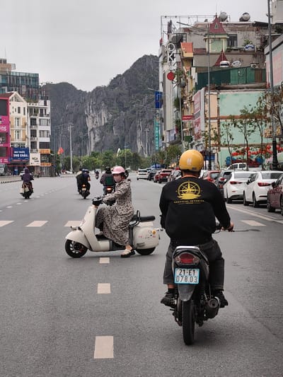 The image shows a street scene in Hong Gai Ward, Vietnam, featuring several people on motorbikes traveling along a multi-lane road. In the foreground, a person with a yellow helmet is riding a motorbike with a license plate that reads "21-E1 078.03". To the left of this person, a woman wearing a pink helmet and a long floral dress is also riding a motorbike. Other motorbikes and cars are visible in both directions of the road. Buildings and mountains can be seen in the background. There are several billboards, trees, and streetlights along the sides of the road.
