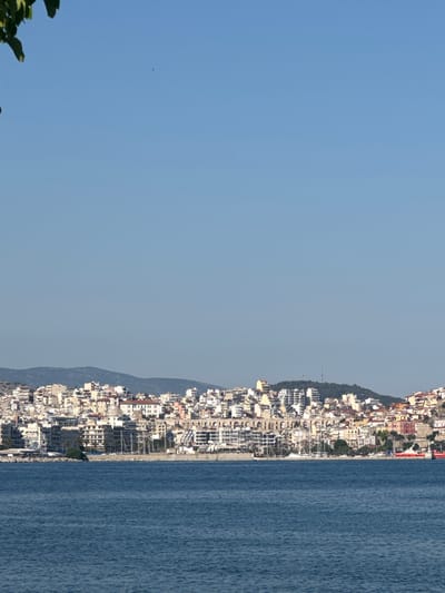 The image shows a panoramic view of the city of Kavala, Greece, from the sea. In the foreground, the deep blue sea is visible with gentle waves. The middle ground displays the coastline of Kavala, with a dense concentration of buildings climbing up a hillside. These buildings are a mix of residential and commercial structures, featuring a variety of architectural styles and colors, with many exhibiting white and light-colored facades. Several boats and yachts are moored along the harbor, and a large red ship is docked to the right. In the background, a range of hills and mountains form a natural backdrop to the city. The sky is clear and blue, indicating a sunny day. There is a hint of foliage from a tree on the left edge of the frame.