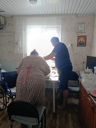 In a room in Каменное, Russia, a man and a woman stand by a table. The woman, wearing a leopard-print dress and an apron, is bent over the table, facing away from the camera. The man, in a dark blue t-shirt and shorts, stands to her right, also facing the table, with his hands near her. A wheelchair is visible to the left of the woman. The room has patterned wallpaper, a ceiling with exposed beams, and a window with lace curtains. A clock on the wall shows the time as approximately 12:07. There is a small framed picture of a religious figure above the clock. Other objects in the room include a television, bread in plastic bags, and a dresser. The floor is made of wood-look laminate.