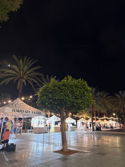 A nighttime outdoor market is depicted in Sant Antoni de Portmany, Spain, with several stalls set up along a paved walkway. In the foreground, a prominent tree stands, with the ground appearing wet and reflective from recent rain, showing a patterned paving design. To the left, a white canopy tent is set up with the words "TEMPORARY TATTOO" displayed on it. A person in a yellow shirt and orange shorts is visible near this stall, operating what appears to be a portable booth setup. Further back, a line of similar white canopy tents stretches into the distance, illuminated by string lights overhead, suggesting a festive atmosphere. Palm trees are visible throughout the scene, indicating a coastal or Mediterranean location. Several people are browsing the stalls or walking between them. The sky above is dark, with some distant lights visible, possibly from buildings or boats.