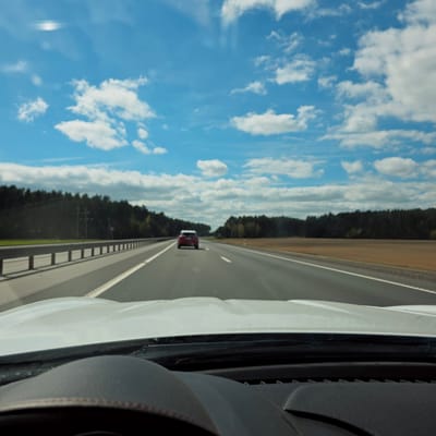This image captures a bright daytime scene from inside a moving vehicle on a highway in Скідзельскі сельскі Савет, Belarus. The perspective is from the passenger side, looking forward and to the left through the window.
On the right side of the frame, parts of the white vehicle's exterior and a dark grey interior element, possibly a dashboard or door panel with textured surfaces, are visible. The main focus is the open road ahead. A multi-lane asphalt highway, delineated by clear white lane markings, stretches into the distance. A metal guardrail runs along the left edge of the road. Further down the highway, a red car is seen, traveling in the same direction.
Beyond the road, a dense, dark green forest forms a consistent tree line along the horizon, suggesting a rural or semi-rural setting. A strip of lighter brown earth or shoulder runs between the highway and the forest. Above, the sky is a vibrant blue, adorned with numerous fluffy white cumulus clouds, indicative of clear and fair weather. Some minor reflections or lens flares are present in the upper sky, likely from the vehicle's window. The overall impression is one of a calm journey through a forested landscape under a sunny sky. No visible text is present in the image.