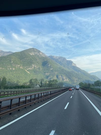 The image shows a view from inside a vehicle, likely a car, driving on a highway. The location appears to be near Dolcè, Italy, judging by the presence of mountains and the type of road. The road is a multi-lane highway with white lane markings and a metal guardrail separating the lanes from a hillside covered with greenery.
There are several vehicles on the road, including what appears to be a red car and a box truck.
The background features a mountain range partially obscured by haze. The sky is blue with scattered white clouds and contrails. The scene conveys a sense of travel and the natural landscape of the region.