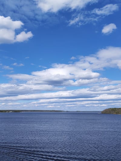 The image depicts a body of water, likely a lake or wide river, under a sky with scattered cumulus clouds. The water is dark blue with slight ripples on the surface. Across the water, a thin strip of land is visible, covered with trees, suggesting a forested shoreline. On the left side of the image, some small structures or buildings are barely discernible on the shoreline. The sky is predominantly blue with a mix of puffy, white clouds and flatter, striated cloud formations.
There are no people visible in the image. No actions or interactions are taking place within the frame. The environment is natural and appears to be a calm, rural setting.
The location given, Irdomatka, Russia, supports the possibility of a lake or river scene within a forested landscape, which is a typical characteristic of Russian geography.