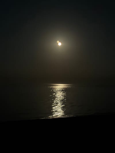 The media file is a photograph taken at night in Çolpon-Ata, Kyrgyzstan. The image is predominantly dark with a low-light effect. In the left half of the frame, two bright, out-of-focus circles of light are visible, suggesting a distant light source such as streetlights or perhaps the moon. The right side of the image features a vertical, dark surface, possibly a wall or a building facade, on which a shimmering, textured reflection of light is cast. This reflection appears to be from a light source, creating an effect reminiscent of water ripples or a textured curtain of light. The overall mood is mysterious and subdued, with the stark contrast between the darkness and the illuminated areas creating a sense of depth and intrigue. The time of day is clearly night, and there are no discernible weather conditions mentioned or depicted. The location cues are limited to the text provided. No people or specific objects are clearly identifiable, and no text is visible within the image itself.