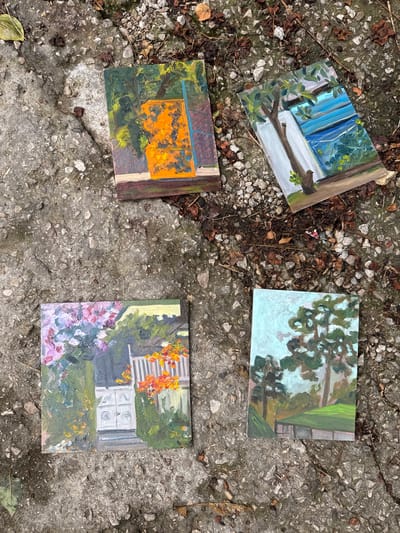 Four small, square paintings are laid out on a concrete surface littered with dried leaves and small stones. The paintings appear to be oil on canvas.

The top left painting depicts a view through a window or doorway, with blue curtains on the left and a brown tree branch extending from the right. Green foliage is visible outside, with dappled sunlight suggesting a daytime scene.

The top right painting shows a cluster of green leaves and branches against a pale blue sky, with a hint of a green structure or fence on the right. The brushstrokes are loose and impressionistic.

The bottom left painting features a vibrant orange rectangular shape, possibly a window or a door, surrounded by dark green and yellow foliage. The background is also rendered in darker tones.

The bottom right painting shows a white building with horizontal siding and some circular details, likely windows or vents. In front of the building, there are bright orange and yellow flowers or foliage, and on the left, there are clusters of pink and purple flowers. The scene appears to be a garden or backyard.

The overall impression is of a series of outdoor scenes, possibly painted en plein air. The concrete surface and scattered leaves suggest an outdoor location, possibly a pathway or courtyard. The lighting in the paintings indicates daylight, with no specific time of day clearly discernible. No people or text are visible.
