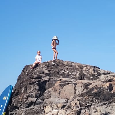 In the city of Alushta, Ukraine, two young girls are pictured standing on a large rock formation. The girl on the left, wearing a white t-shirt and shorts, sits on the edge of the rock, looking towards the viewer. The girl on the right, wearing a patterned hat and bikini bottoms, stands on a higher point of the rock, with her hand near her face. A blue paddleboard is visible on the left side of the frame, leaning against the rock formation. The background is a clear, bright blue sky.