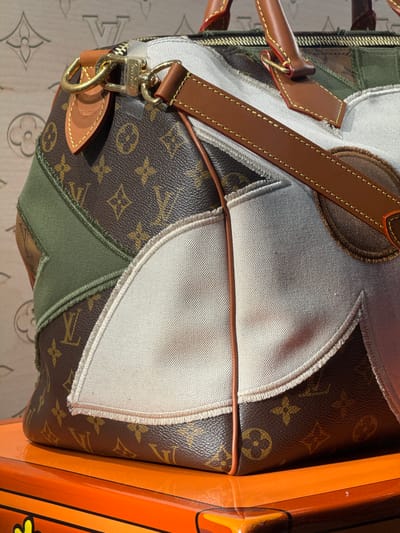 A close-up, eye-level shot captures a Louis Vuitton Speedy bag with a patchwork design. The bag is made of the iconic brown Louis Vuitton monogram canvas, interspersed with panels of white canvas, olive green fabric, and brown denim. The bag features brown leather handles and trim, with gold-toned hardware. A Louis Vuitton tag hangs from one of the handles. The bag rests on an orange surface with yellow accents, possibly a piece of furniture. The background appears to be a patterned surface, possibly wallpaper, with a faint Louis Vuitton motif. The image is well-lit, with the light source coming from the top right. The scene is indoors, with no discernible city or specific location information available within the image itself.