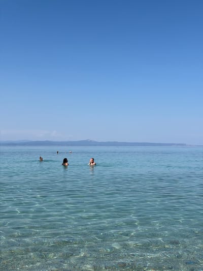 This media file depicts a tranquil scene of people swimming in the clear, turquoise waters of Kassandra, Greece. The image is oriented vertically, showcasing a vast expanse of bright blue sky on the left, transitioning to the shimmering sea on the right. The water is calm, with gentle ripples reflecting the sunlight, creating a mesmerizing pattern on the surface. The seabed is visible in shallower areas, hinting at a rocky or sandy bottom.
Several individuals are scattered across the water, enjoying a swim. In the foreground, a person is partially submerged, their head and shoulders visible above the surface. Further out, another swimmer is seen with their head turned towards the viewer, a faint smile suggested by their expression. Other figures are visible in the distance, also enjoying the refreshing water. The background features a hazy, mountainous coastline, characteristic of the Mediterranean landscape. The lighting suggests it is a bright, sunny day, likely late morning or early afternoon, given the clear blue sky and the intensity of the sunlight on the water. The overall atmosphere is one of relaxation and leisurely enjoyment of a summer day by the sea. No text is visible in the image.
