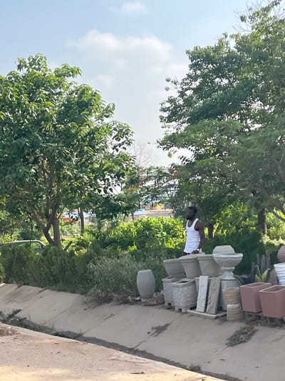 The image shows an outdoor scene in what appears to be a residential area of Kuchingoro, Nigeria. A man wearing a white tank top is visible, standing next to a stack of concrete and terracotta pots and other decorative concrete items, presumably for sale or display. The environment is green with trees and other plants. A sidewalk or paved area is adjacent to the pots. The sky is partially visible with some clouds.
