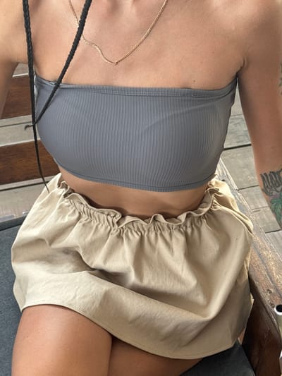 The image shows a close-up, top-down view of a person seated, likely in an outdoor or semi-outdoor setting within Hà Giang 1 Ward, Vietnam. The subject, visible from the upper chest to the upper thighs, wears a grey ribbed crop top and a light beige or khaki-colored skirt with a ruffled, elasticated waist. A thin gold chain necklace is visible around their neck, and strands of dark braided hair extend across their chest. A tattoo featuring green and black ink, possibly depicting foliage or an abstract design, is visible on their upper right arm.
The person is seated on a grey surface, possibly a cushion or upholstered chair. Behind and to the side of the subject, a wooden structure, likely a table or bench, with visible grain and fasteners, suggests a rustic or natural environment. The bright, even lighting indicates it is daytime. The activity captured is simply sitting. No visible text is present in the image.