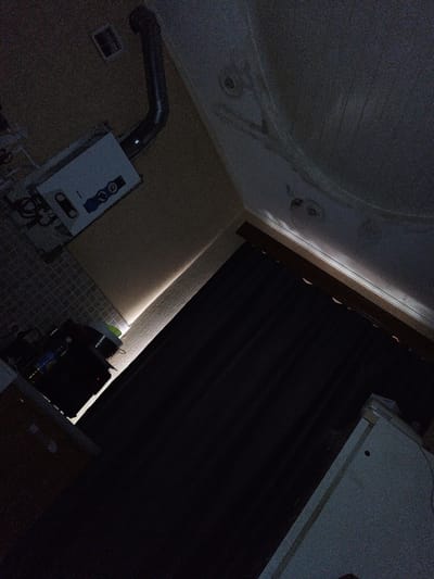 The image depicts an interior scene, likely a room or hallway, oriented such that the ceiling occupies a significant portion of the upper frame. The lighting is dim, with a bright strip of light illuminating the junction between the wall and the ceiling in the mid-ground.
In the upper left of the image, a white gas water heater is mounted on a wall. A metal vent pipe extends from the heater upwards and curves towards the ceiling. Below the water heater, a tiled surface is visible, and on it sits a dark appliance, possibly a stove or oven, with some items on top.
The right side of the image is dominated by a dark curtain that extends from near the ceiling down to the bottom of the frame. Above the curtain, the ceiling is textured and appears to have some circular fixtures or stains.
The bottom right corner shows a portion of a light-colored appliance or cabinet, with a dark object on top.
The image is grainy, suggesting it was taken in low light conditions. The overall impression is of a functional but somewhat cluttered indoor space. The location is identified as Beloretsk, Russia.